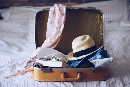 An open suitcase is on a bed, filled with travel essentials. Items include a straw hat, sunglasses, a floral scarf, denim clothes, a book, and a passport. The suitcase is beige with a quilted interior, set against a striped bedspread.