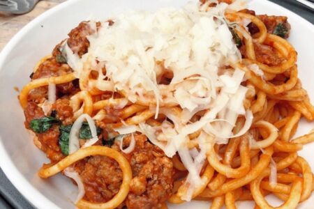 A plate of spaghetti topped with a meaty tomato sauce, fresh basil, and a generous sprinkle of grated cheese. The dish is served in a white bowl on a table setting with cutlery and a napkin nearby.