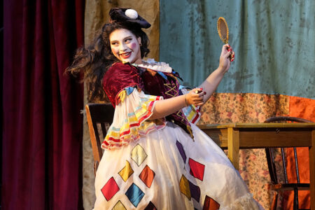 A performer in a colorful costume and feathered hat holds a handheld mirror, smiling on stage. The dress features multicolored diamond patterns, and there's a table and chair in the background.