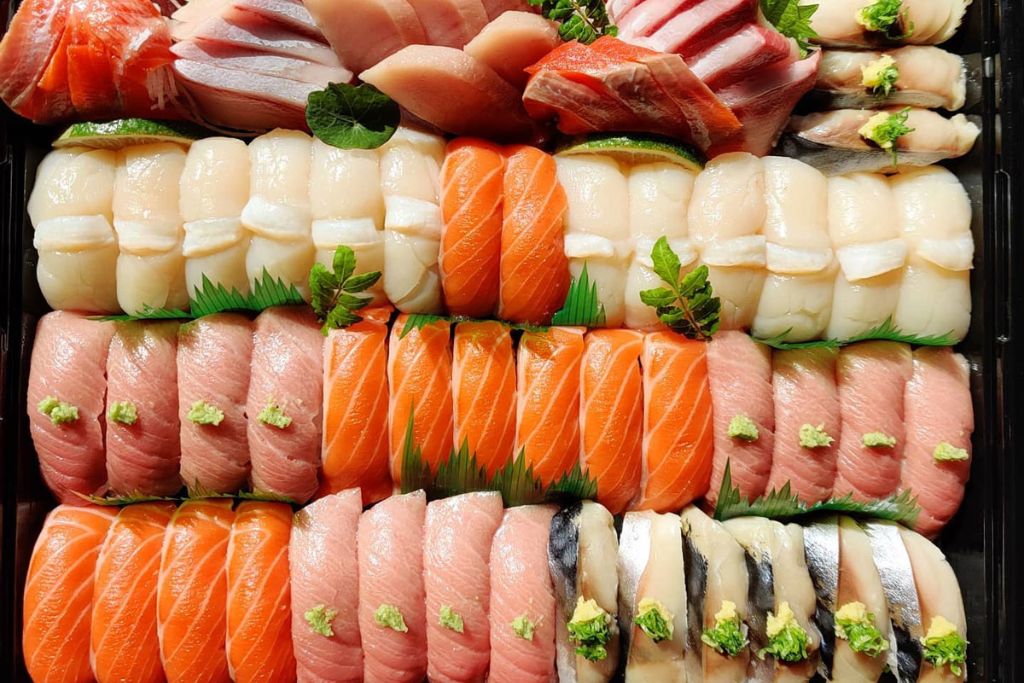 An assorted tray of sushi with rows of various nigiri, including tuna, salmon, white fish, mackerel, and scallops, each topped with a small amount of wasabi and garnished with green leaves.
