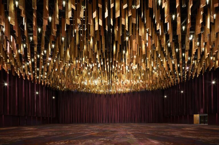 A large, empty hall features an elaborately designed ceiling with numerous wooden panels hanging like stalactites, illuminated by small lights. The walls and floor are adorned in rich, dark tones, creating a warm, elegant atmosphere.