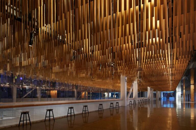 A modern interior with a ceiling adorned with numerous vertical wooden panels creating a wave-like pattern. Tall windows on the left offer a view, while a row of stools lines the spacious wooden floor below.