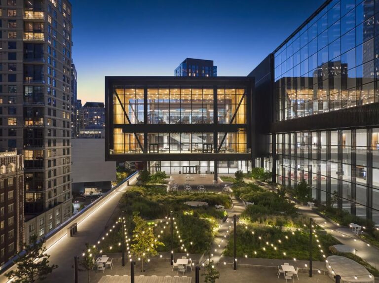 A modern, illuminated office building with a rooftop garden and string lights. The garden features seating areas and pathways surrounded by greenery. Skyscrapers are visible in the background against a twilight sky.