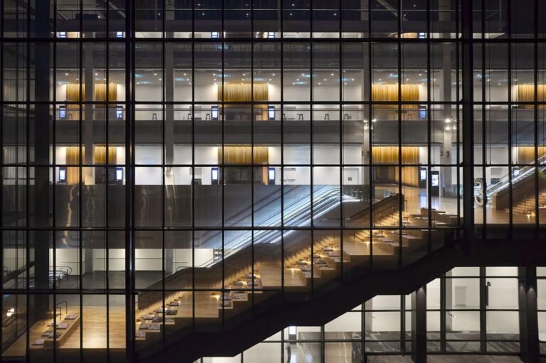 A modern, glass-walled interior with illuminated staircases and seating areas. The design features multiple levels, with warm lighting and wooden accents creating a sleek, contemporary atmosphere.