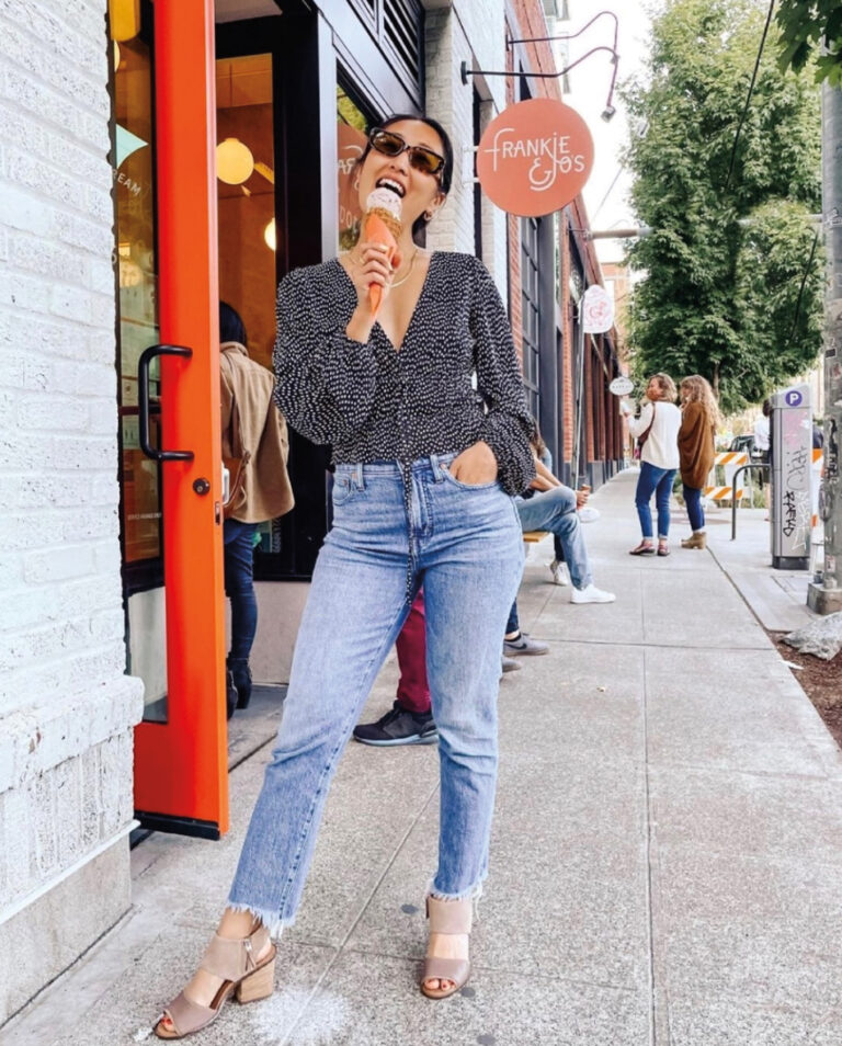 A person enjoying an ice cream cone stands outside a shop called "Frankie & Jo's." They're wearing sunglasses, a patterned long-sleeve shirt, and jeans. People are walking on the sidewalk, and there's greenery in the background.