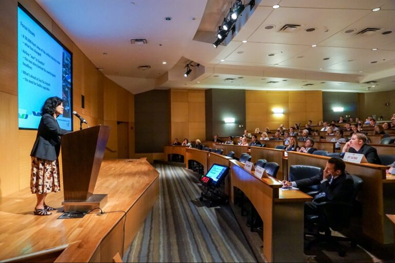 A speaker is presenting at a podium in a modern lecture hall. The audience is seated in tiered rows, attentively watching the presentation. A large screen displays text and images. The room is well-lit with wood-paneling and a striped carpet.