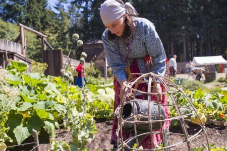 A person in historical clothing waters plants in a garden using a watering can. The garden is lush, with various plants and a wooden structure around it. People and an old-style building are visible in the background under a clear blue sky.