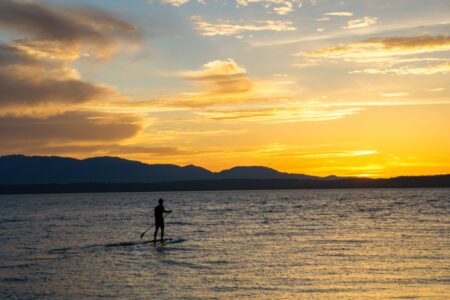 Silhouette of a person paddleboarding on a calm sea during sunset. The sky is filled with orange and yellow hues, and distant mountains are visible on the horizon.