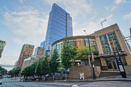 Urban scene featuring a tall glass skyscraper, surrounded by modern buildings and trees. Below, there's an entrance to a public transit station on a slightly inclined street under a partly cloudy sky.