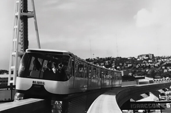 Black and white photo of a monorail traveling on an elevated track. In the background, there's a tall structure and a hillside with buildings. The image captures a cityscape with a blend of modern and urban elements.