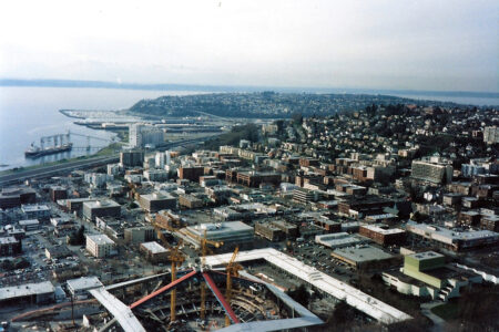 Aerial view of a coastal city with a dense cluster of buildings, roads, and a construction site with cranes in the foreground. The coastline and distant hills are visible under a cloudy sky.