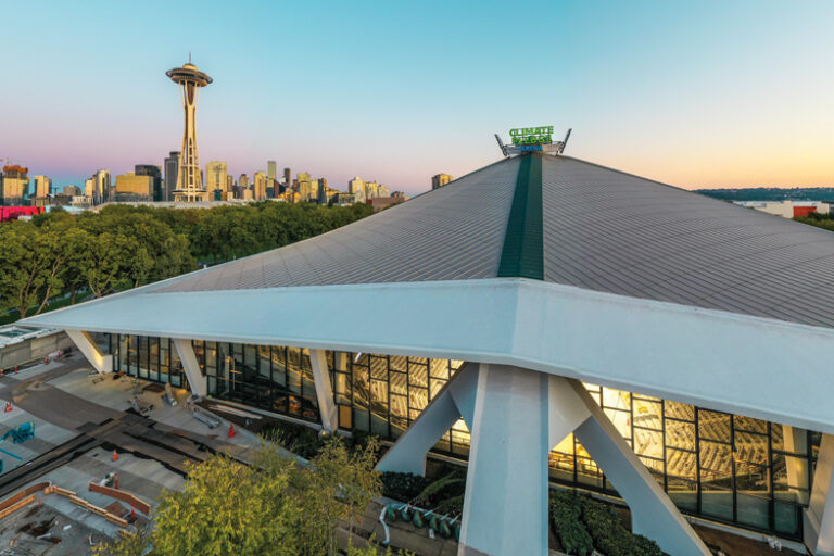 Aerial view of the Climate Pledge Arena in Seattle, with the iconic Space Needle visible in the background. The arena's modern design contrasts with the city skyline at sunset, under a clear blue and pink-hued sky.