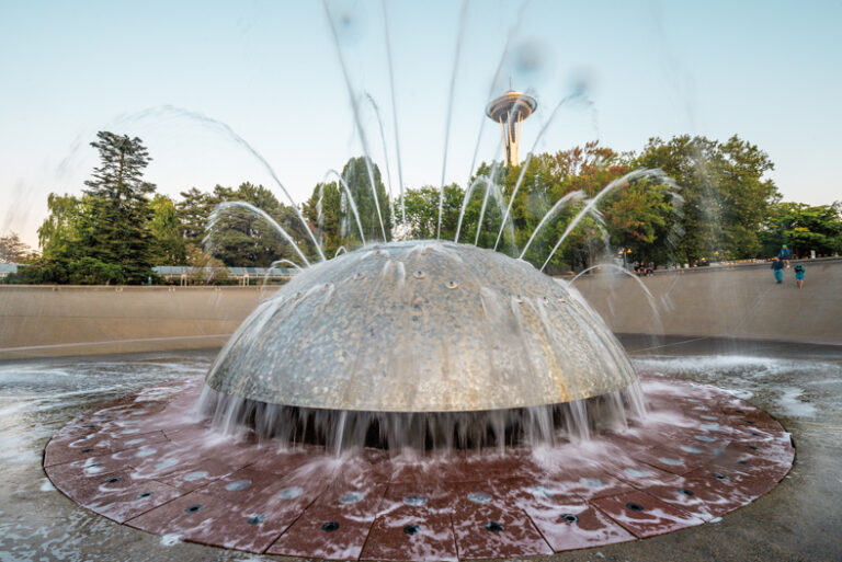 A large, spherical fountain sprays water in an outdoor plaza. Trees and the top of a tall needle-shaped tower are visible in the background under a clear sky.
