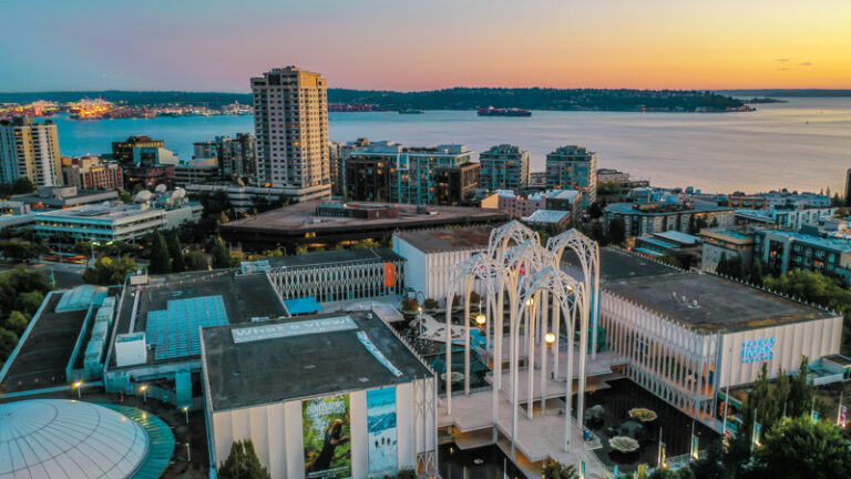 Aerial view of a cityscape at sunset, featuring modern architecture with tall, arching structures. In the background, a body of water and more urban buildings are visible under a colorful sky.