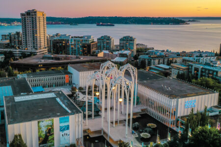 Aerial view of a cityscape at sunset, featuring modern architecture with tall, arching structures. In the background, a body of water and more urban buildings are visible under a colorful sky.