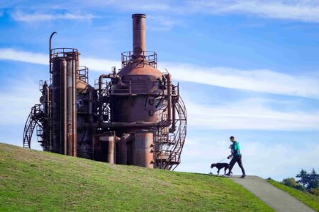 A person walks a dog along a grassy hill near large, rusted industrial structures.