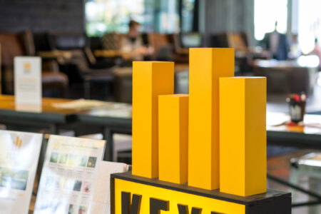 A modern lobby featuring a yellow and black KEXP sign on a wooden counter. The background has seating arrangements, people sitting, and decorative elements, creating a relaxed atmosphere.