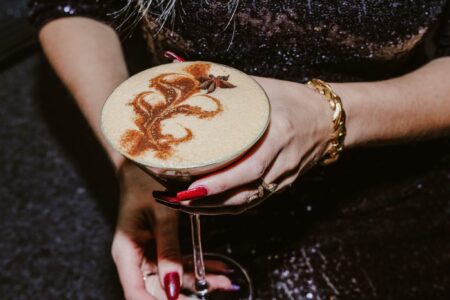 A person with red-painted nails holds a cocktail glass filled with a frothy, tan beverage. The drink surface is adorned with intricate brown swirls and a star anise garnish. The person wears a sparkly outfit and gold jewelry.