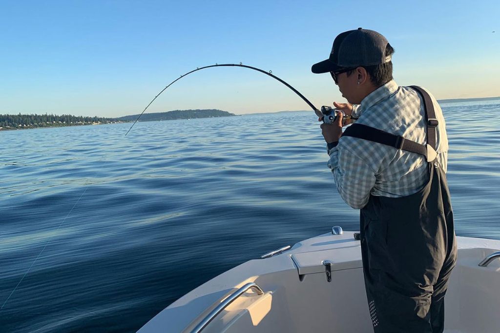 A person wearing a cap and checkered shirt stands at the bow of a boat, holding a fishing rod with a bent line over calm blue water on a sunny day.
