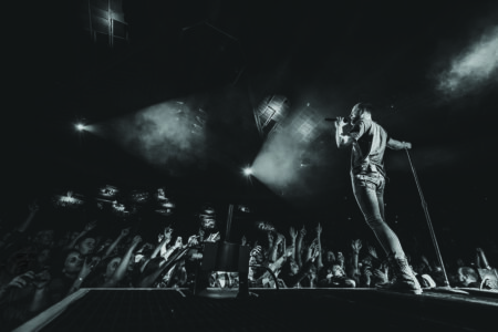 A black-and-white photo of a singer performing energetically on stage with a microphone, facing a cheering audience. Stage lights create dramatic shadows, and the crowd reaches towards the stage, capturing the excitement of the concert atmosphere.