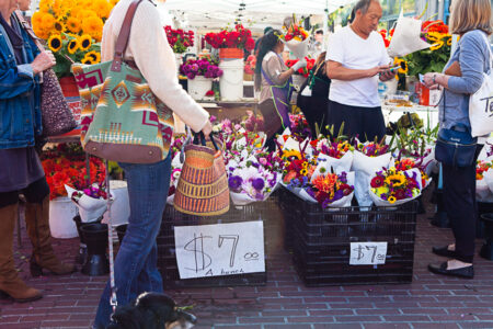 People browse a vibrant flower market, with colorful bouquets in black crates selling for $7 each. Sunflowers and other flowers are displayed in buckets. Some shoppers carry tote bags and baskets while a dog is partially visible among them.