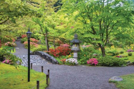 A tranquil garden scene featuring a stone path with a small bridge, surrounded by lush green trees and colorful blooming flowers. A traditional stone lantern and a black lamppost are situated along the path, adding to the serene atmosphere.