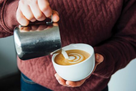 A person in a maroon sweater is pouring steamed milk into a cup of coffee, creating a latte art design. The cup is held in one hand, and the milk is poured from a metallic pitcher.
