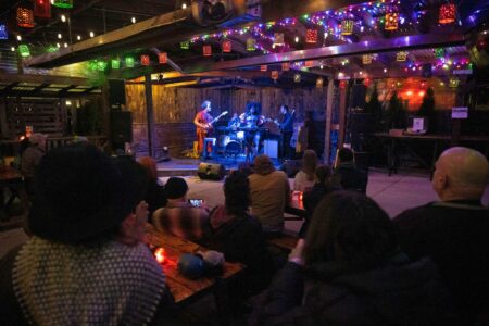 A band performs on an indoor stage adorned with colorful string lights. The audience sits at wooden tables, some capturing the moment on their phones. The cozy venue has a warm, inviting atmosphere with dim lighting.