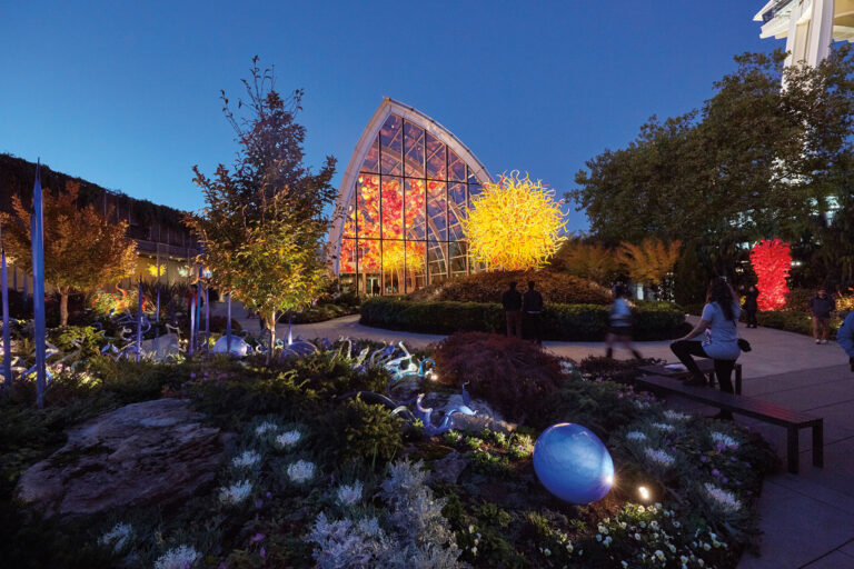 A vibrant evening scene at a botanical garden, showcasing a large glass greenhouse with colorful illuminated art inside. Surrounding the greenhouse are lush plants, trees, and sculptures, with several people enjoying the illuminated display.