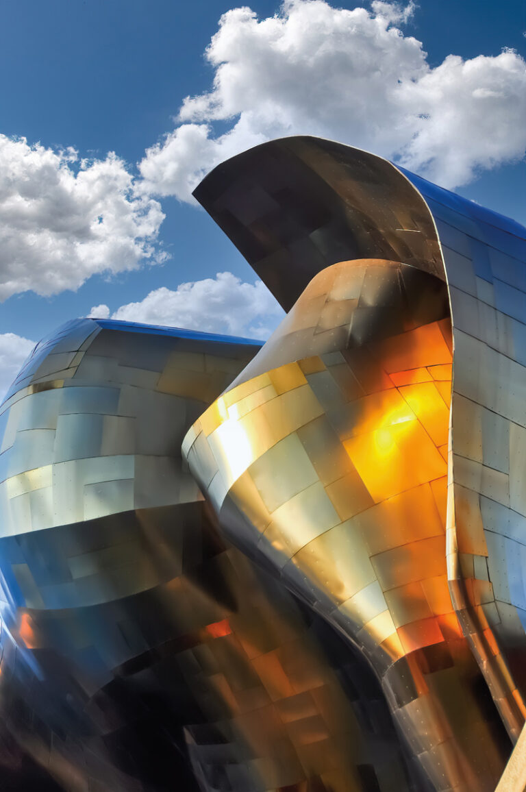 A close-up view of a futuristic building with reflective, metallic surfaces in gold, bronze, and silver tones. The curving structure is set against a blue sky with scattered fluffy clouds, creating an abstract, dynamic appearance.