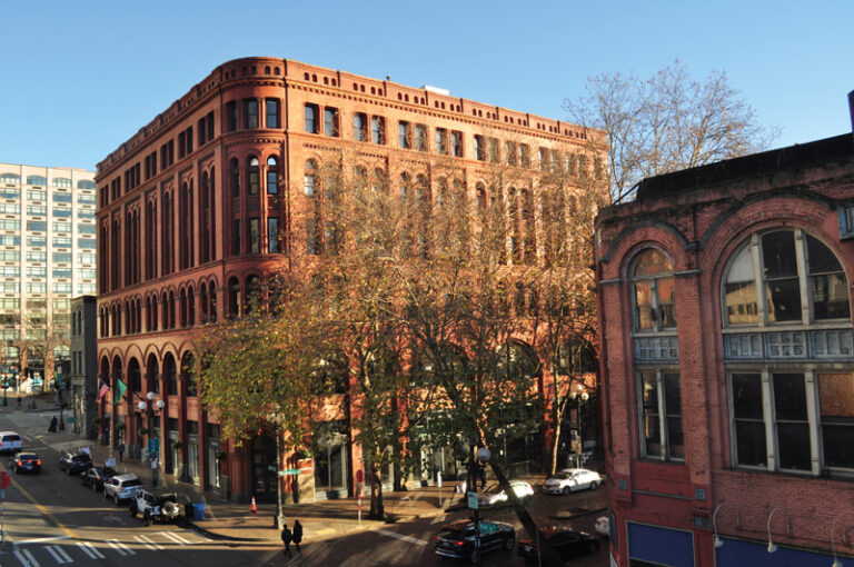 A large, historic red-brick building with arched windows stands on a city corner, surrounded by trees. Cars and pedestrians move through the intersection on a sunny day. A modern building is visible in the background.