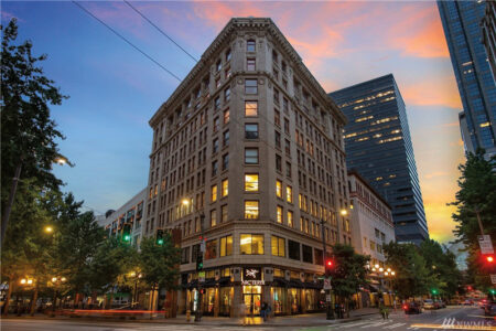 A historical multi-story building with ornate architecture stands at a street corner during sunset. The sky displays colorful clouds, and the street is lined with trees and modern tall buildings. Illuminated streetlights and storefront windows brighten the scene.