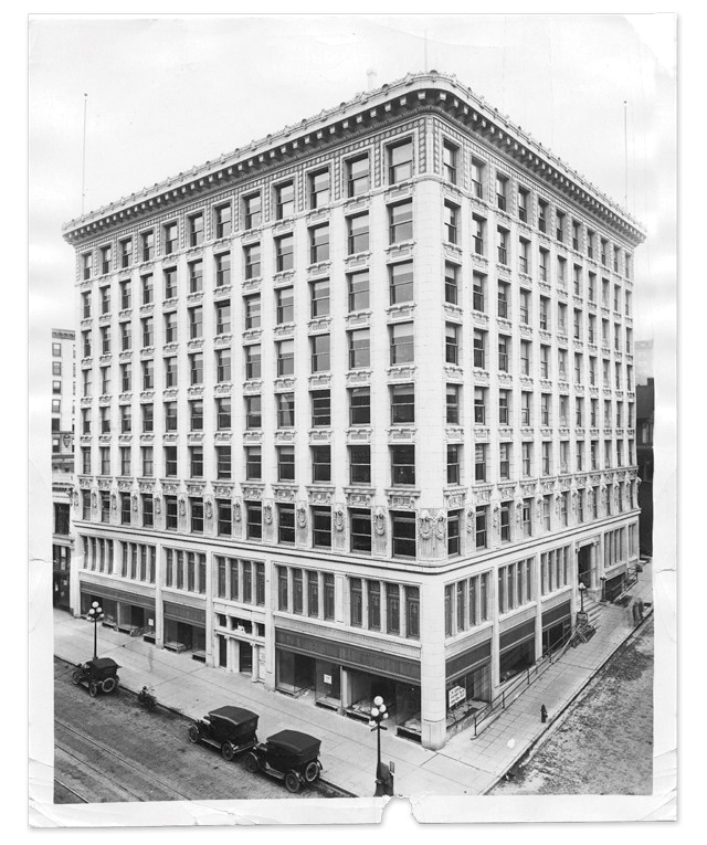 Black and white photo of a multi-story historic building on a corner street, with early 20th-century cars parked along the sidewalk. The architecture features decorative columns and cornices. The streets are visibly empty.