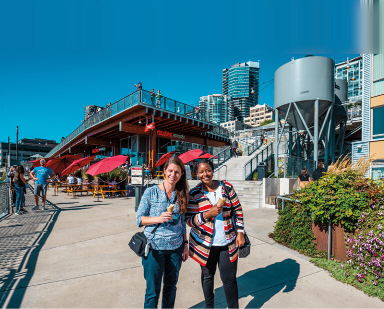 Two women smiling and holding ice cream cones stand on a sunny sidewalk. Behind them are a modern building with red umbrellas and a staircase. The sky is clear and blue, and leafy plants are visible on the right.
