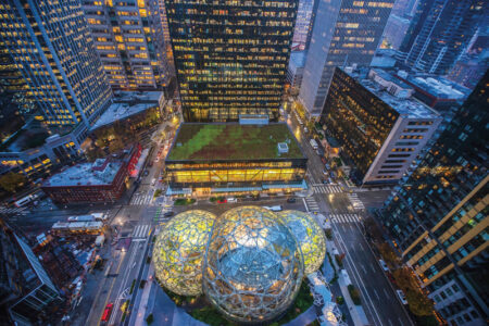 Aerial view of a city intersection at dusk, featuring three spherical glass structures with geometric patterns on a corner. Surrounding buildings are illuminated, and traffic is visible on the streets. One building has a green rooftop garden.