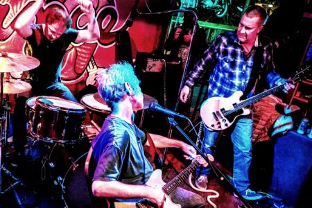 A lively band performance in a colorful venue. A seated guitarist and singer faces a standing bassist. In the background, a drummer energetically plays in front of a vibrant mural on the wall.