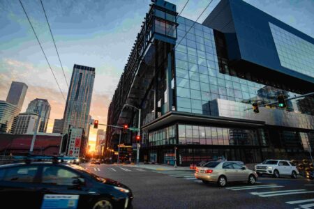 Cars drive through a city intersection at sunset, with tall modern glass buildings reflecting the sky.