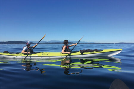 Two people in a green kayak in the glassy blue water of Puget Sound. A small sliver of land is on the horizon. A deep blue sky fills the background.