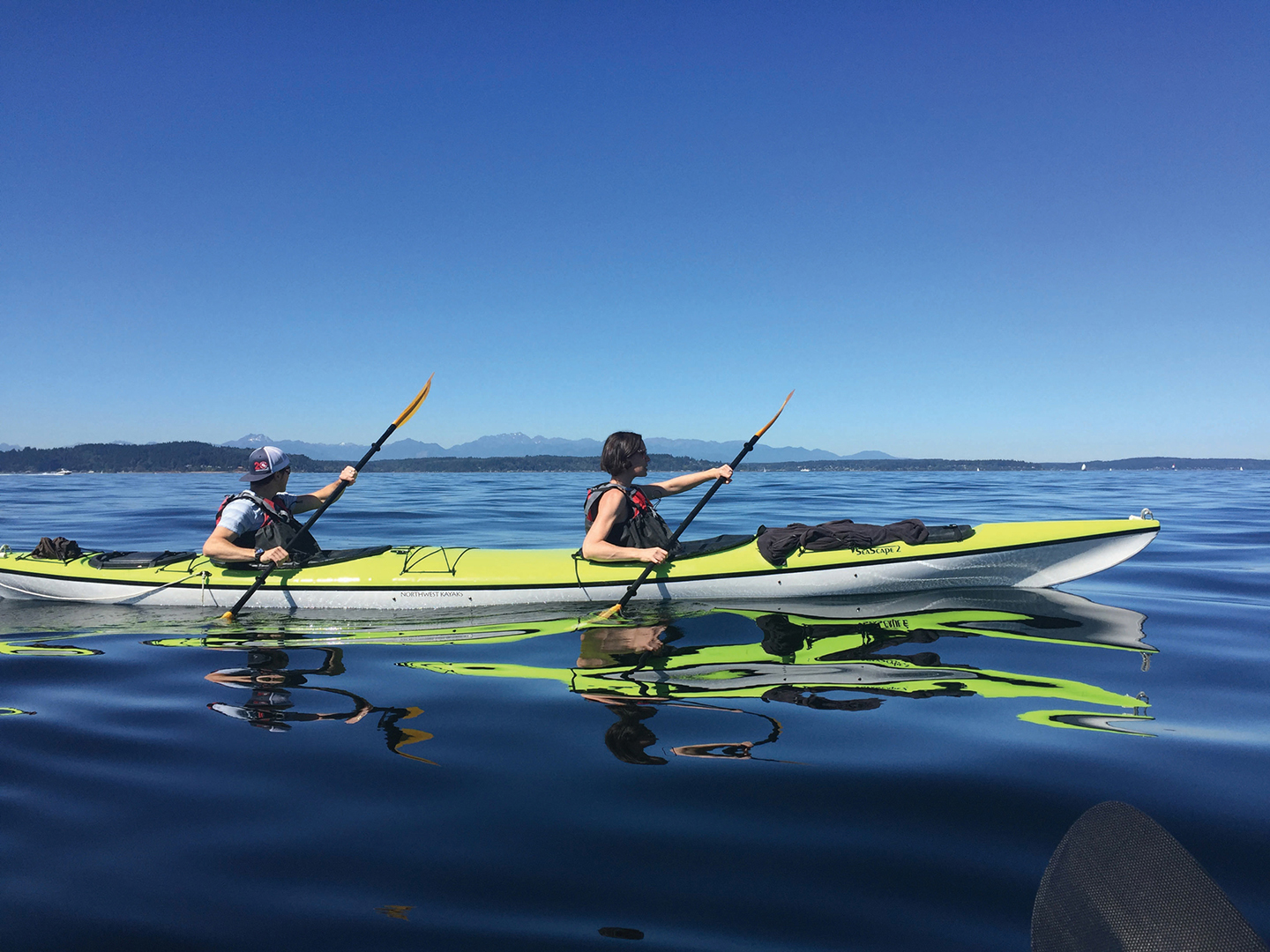 Two people in a green kayak in the glassy blue water of Puget Sound. A small sliver of land is on the horizon. A deep blue sky fills the background.