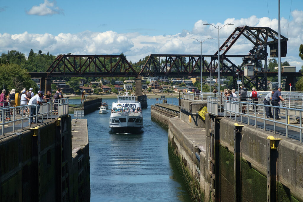 A white Argosy Cruises ship is aligned with the opening of the Ballard Locks as it moves forward into position. People stand on either side of the locks' opening, watching the boat traffic. A rusted metal train bridge crosses the horizon in the background.
