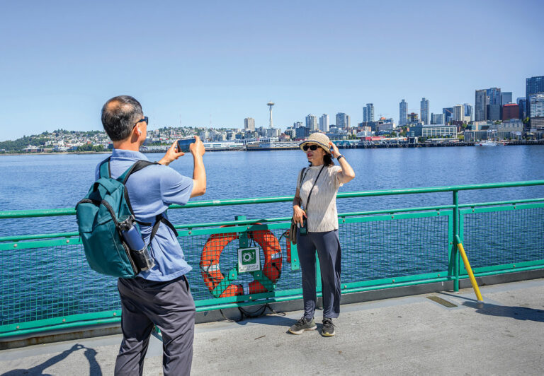 A man in a blue polo with a blue backpack takes a photo of a woman wearing a tan bucket hat, a cream shirts, and blue pants standing on the deck of a ferry. The ferry has a green metal railing. The blue waters of Elliott Bay and the Seattle skyline are seen in the background, including the Space Needle in the middle of the image.