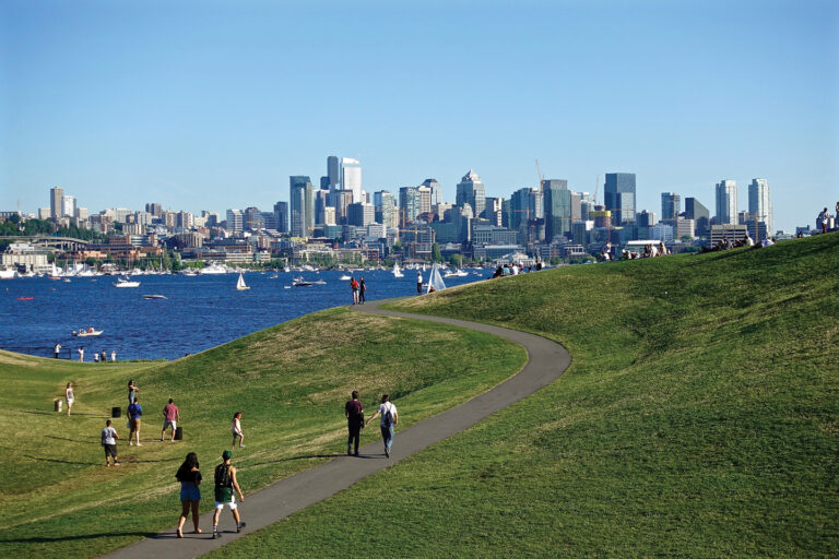 A rolling green hill with a winding cement pathway fill the foreground. People in shorts, t-shirts, and pants walk along the pathway and hill. The blue water of Lake Union is in the middle ground and is dotted with white motorboats and sailboats. The Seattle skyline is in the background set against a light blue sky.
