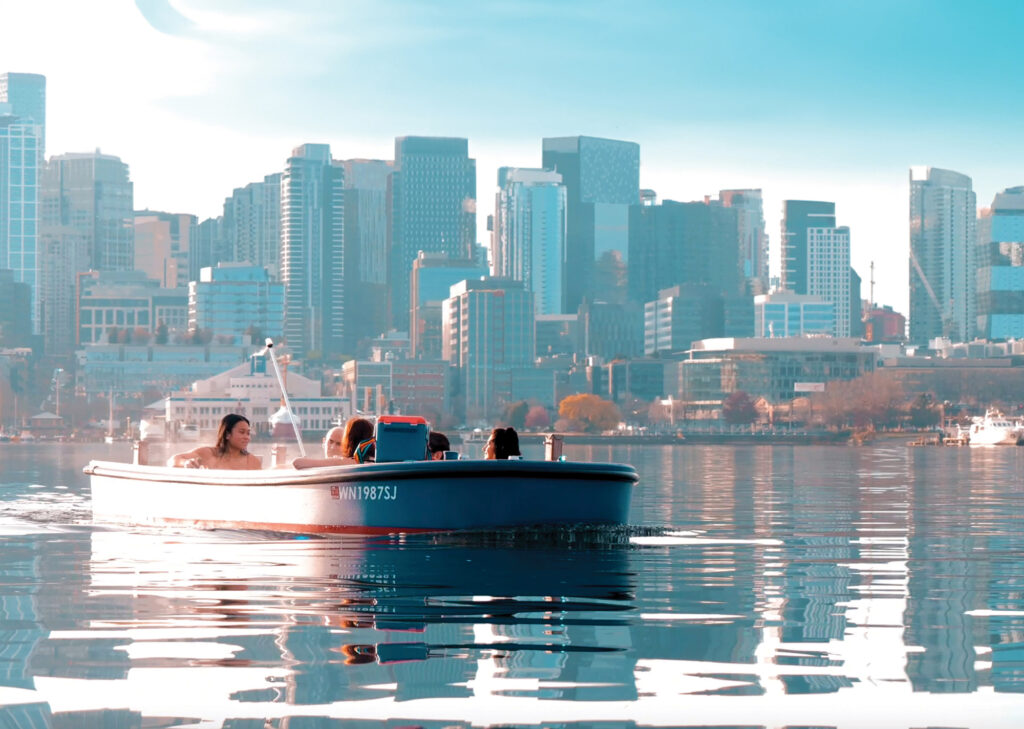 A group of 5 people cruise in a blue hot tub boat on the glassy waters of Lake Union. Skyscrapers fill the background and a dream-like haze filters the image.
