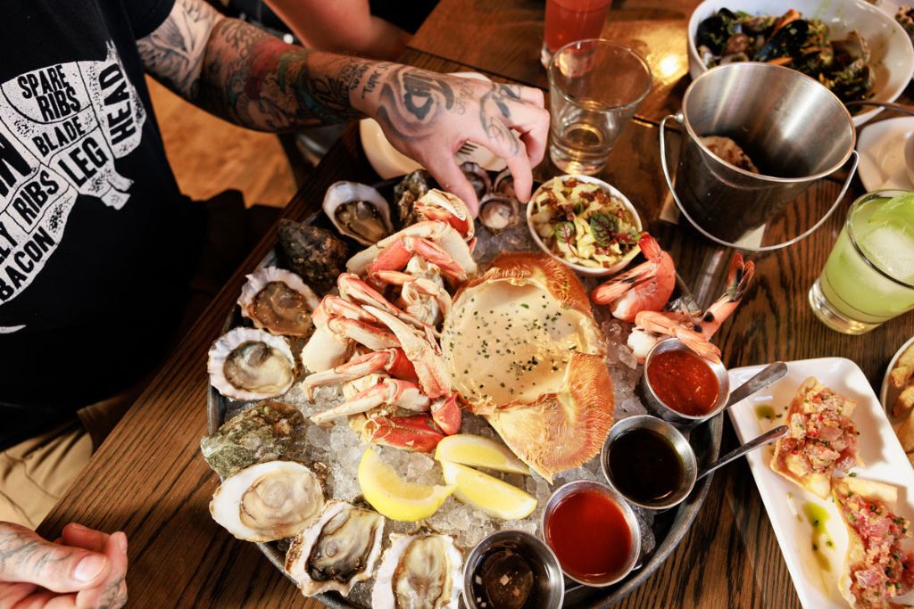 A person with tattooed arms sits at a table, reaching for a platter of assorted seafood including oysters, crab legs, shrimp, lemon wedges, and dipping sauces, with drinks and side dishes nearby.