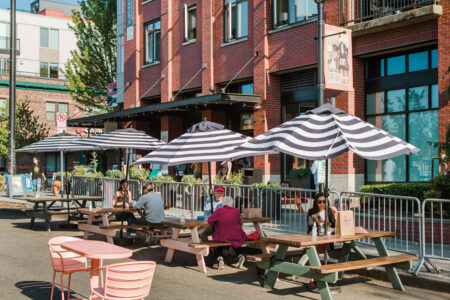 People sit at outdoor tables under black-and-white striped umbrellas near a red brick building. The area is enclosed by metal barriers. The scene is sunny, with a small tree and additional tables visible in the background.