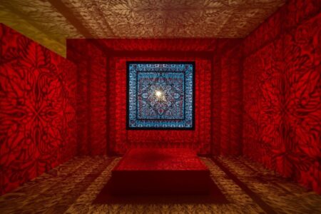 A room illuminated with intricate, symmetrical patterns in red and blue, projected from a central light source. The geometric designs cover the walls, ceiling, floor, and a raised platform in the center.