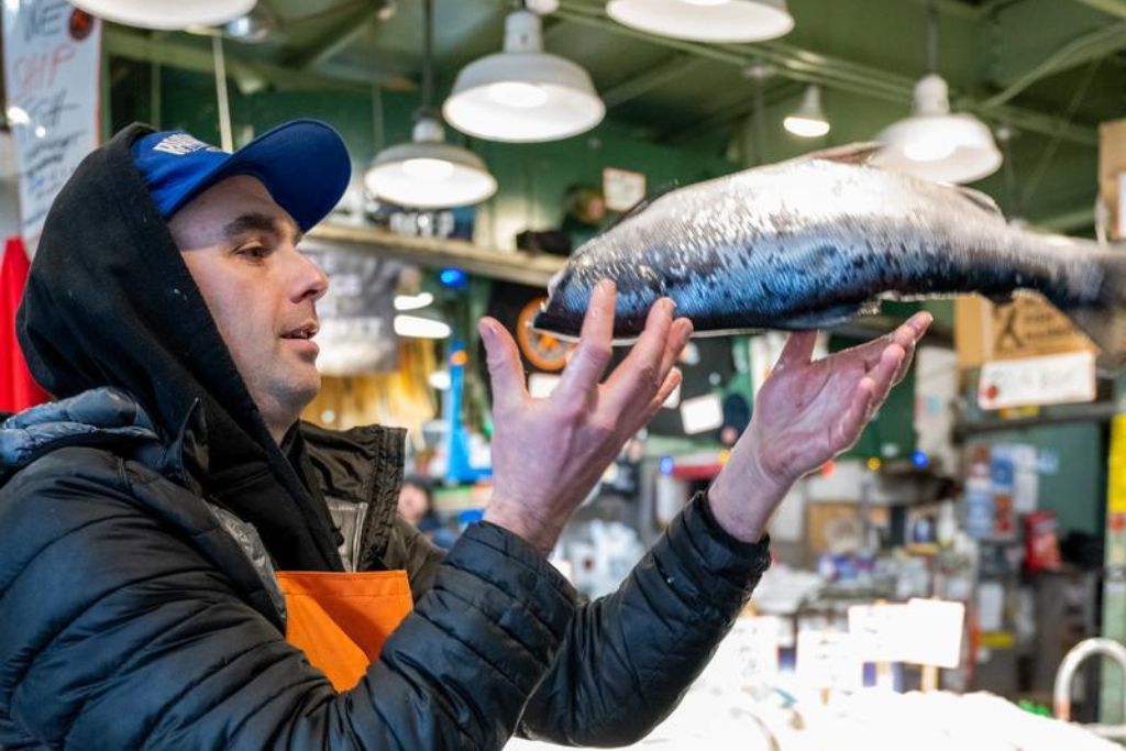 A person wearing a blue cap and black jacket tosses a large fish in the air inside a busy indoor market with bright lighting overhead.