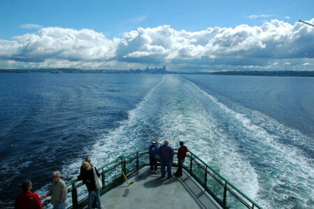 A ferry moves away from the city skyline under a cloudy sky, leaving a wake in the water. Several people stand on the deck, enjoying the view of the ocean and distant cityscape.