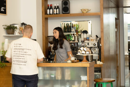 An Asian American woman standing behind a beverage counter, taking an order from a man in a white t-shirt with his back to the camera.