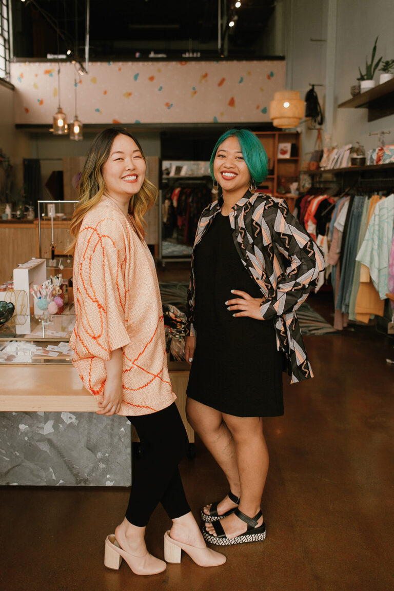 Two Asian women, one with dark hair and highlights wearing pink and the other with emerald green hair wearing black, stand facing each other, heads turned and smiling at the camera, in